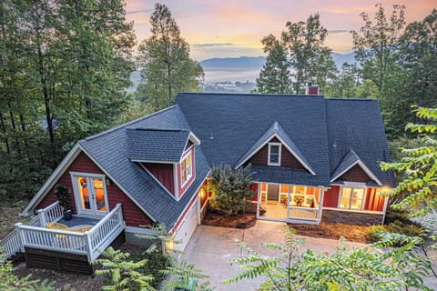 A charming mountain home nestled among lush trees, featuring a red and brown exterior, a welcoming front porch, and a private deck. The property overlooks a stunning valley with misty mountains in the background, illuminated by a beautiful sunset.