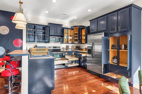 Featuring sleek navy cabinets with gold hardware, ample storage with pull-out drawers, and modern stainless steel appliances, it’s a chef’s dream. Open shelving showcases elegant glassware, while a unique accent wall and bold red barstools add a touch of personality. Perfect for cooking, entertaining, and making lasting memories.