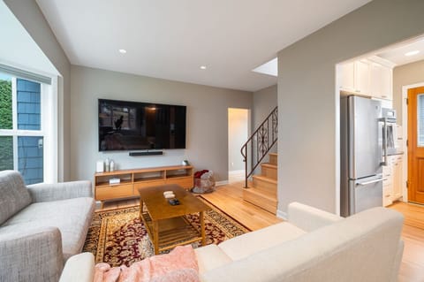 MAIN FLOOR - LIVING ROOM: The inviting living room boasts large windows with lush views, a light gray sofa set for 2 people, a navy velvet love seat, and a Persian rug beneath a wooden coffee table.