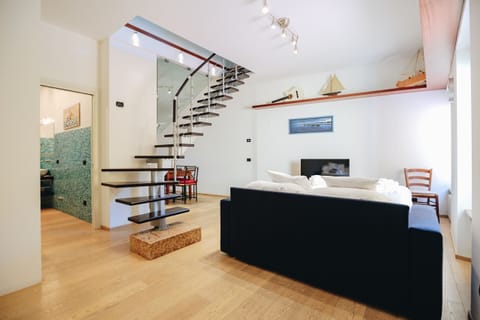 Elegant living room with modern wood and steel staircase, part of a refined two-level apartment. The bright and well-maintained space combines contemporary style with decorative touches inspired by the lake, such as model boats and natural tones. The relaxation area features a comfortable sofa, TV, and direct access to the upper sleeping area, creating a welcoming and functional atmosphere.