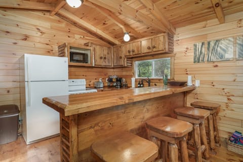 Rustic cabin kitchen with a polished wooden breakfast bar and matching stools. Cozy space with natural wood walls, white appliances, and charming forest artwork for a warm, inviting atmosphere 🌲🍳🪵