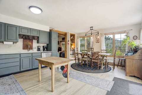 Charming open kitchen and dining space with sage green cabinetry, wood accents, and large windows that fill the room with natural light and forested views.