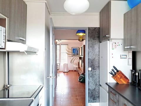 Cuisine compacte avec meubles en bois clair, plaque de cuisson, réfrigérateur et accessoires modernes. Carrelage au sol et ouverture vers une autre pièce.