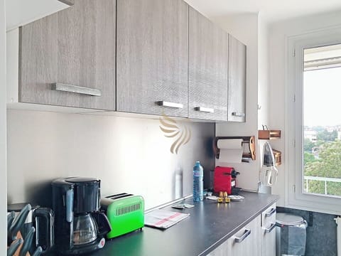 Cuisine lumineuse avec placards en bois, machine à café, grille-pain vert et vue dégagée par une grande fenêtre. Essuie-tout et bouteilles d'eau sur le plan de travail.