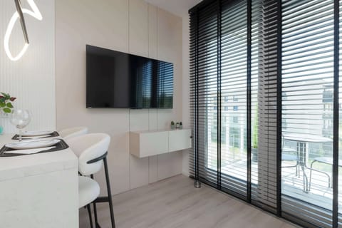 A modern, minimalist studio featuring a large wall-mounted TV, a sleek shelf with decorative items, and spacious floor-to-ceiling windows with black blinds that let in natural light, creating a cozy and stylish space for relaxation.