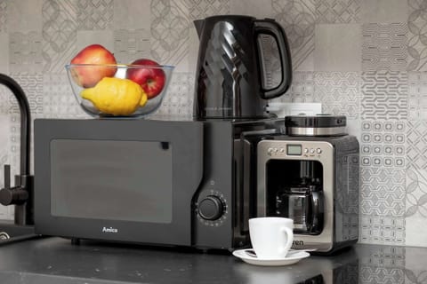 A close-up of a modern kitchen countertop with a microwave, espresso machine, and fruit bowl. Sleek black appliances contrast with the clean white background.