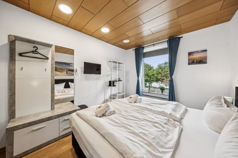 Modernes Doppelzimmer mit Boxspringbett, Holzdecke, TV, offener Regalwand und Fenster mit Vorhängen – hell, gemütlich und ideal für den Aufenthalt.