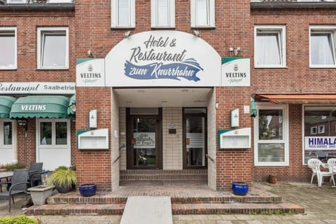 Backsteinfassade mit überdachtem Hoteleingang und Restaurant „Zum Knurrhahn“ – zentraler, gut sichtbarer Zugang für Gäste.