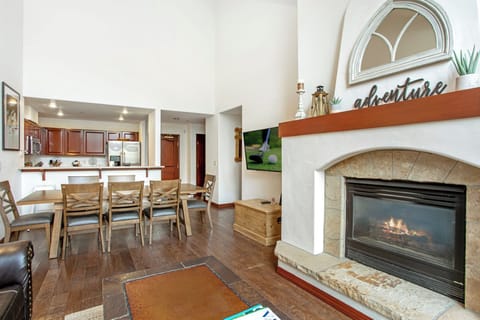 Spacious living area with a large dining table, modern kitchen, and a cozy stone fireplace. The wide-angle view showcases high ceilings and a wall-mounted TV in the background.