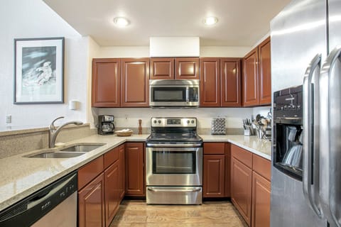 Modern kitchen with stainless steel appliances, brown cabinets, granite countertops, wood flooring, and a double sink. Refrigerator, oven, and microwave are included. Wall art is present in the background.