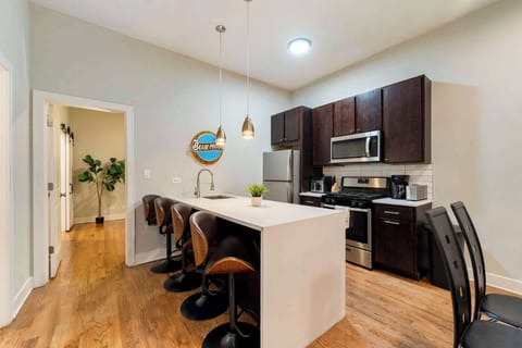 A modern kitchen with dark cabinets, stainless steel appliances, a white island, and bar stools on wooden flooring.
