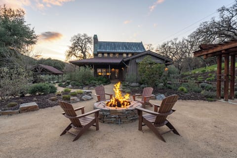 This gorgeous property features a central stone fire pit with a roaring fire, perfectly complemented by rustic Adirondack seating. The dark wood home, glowing with interior warmth, provides an intimate backdrop for evening relaxation.