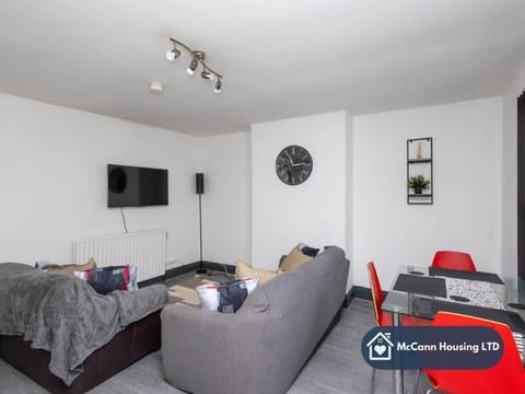 A cozy, modern living room with a grey sofa, wall-mounted TV, and a dining table with red chairs. The space is bright and inviting, ideal for relaxing or entertaining in a comfortable home setting.
