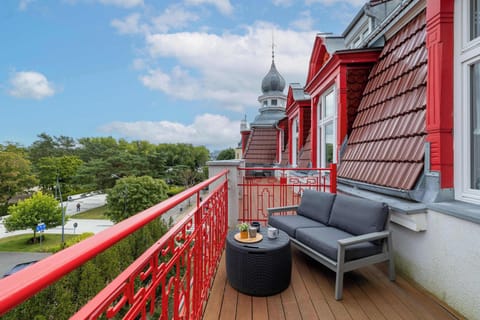A red balcony with outdoor chairs and a small table. The view overlooks greenery and neighboring houses.