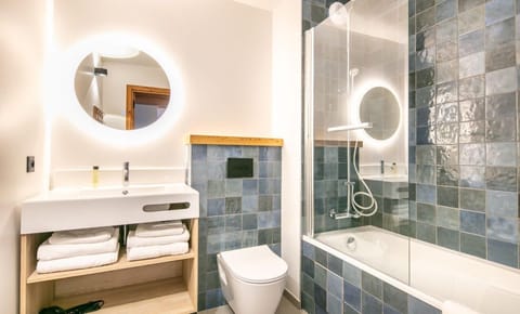 A modern bathroom featuring a stylish circular mirror, sleek sink, and tiled walls in calming blue tones, perfect for relaxation after a day in the Alps.