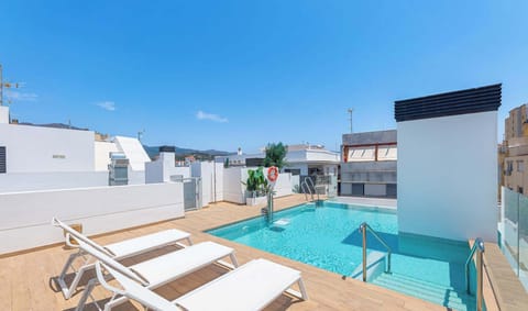 Rooftop swimming pool with crystal clear water, wood-look decking, and multiple white sun loungers. Glass safety barriers, tropical plants, and views over surrounding buildings under blue sky.