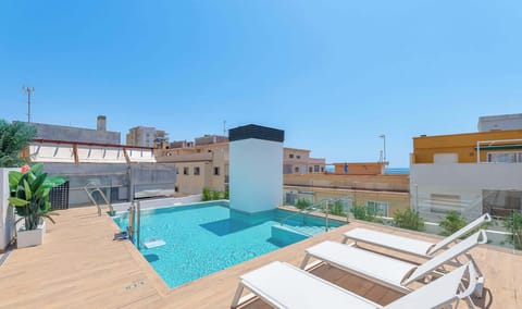 Rooftop swimming pool with crystal clear water, wood-look decking, and multiple white sun loungers. Glass safety barriers, tropical plants, and views over surrounding buildings under blue sky.