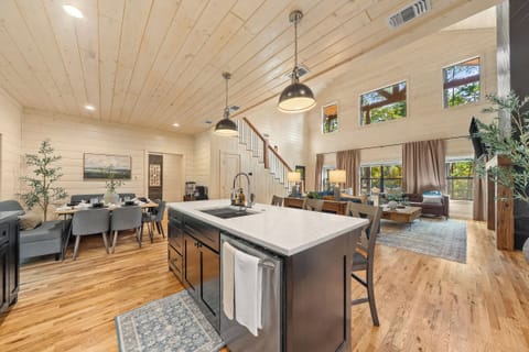 Bright great room where kitchen, dining, and living come together under soaring ceilings and tree framed windows.