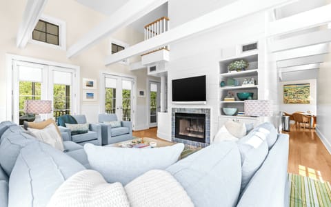 Relax in your spacious living room, where soaring ceilings and soft blue sofas invite you to unwind by the fireplace beneath a wall-mounted TV, with natural light streaming through tall windows.