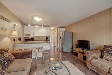 A living room with a couch, armchair, glass coffee table, and wall-mounted TV. The open kitchen has white cabinets, stainless steel appliances, and a breakfast bar with stools.