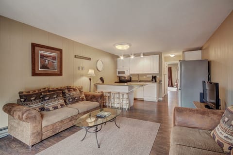 A cozy living room with a brown sofa, patterned pillows, a glass coffee table, and a TV. It is adjacent to a kitchen area with white cabinets, an island with two stools, and various appliances.