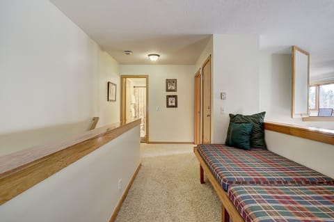 A hallway with carpeted flooring, wooden trim, and a ceiling light leads to various rooms. A cushioned bench with green and plaid pillows is placed along a wall on the right side.