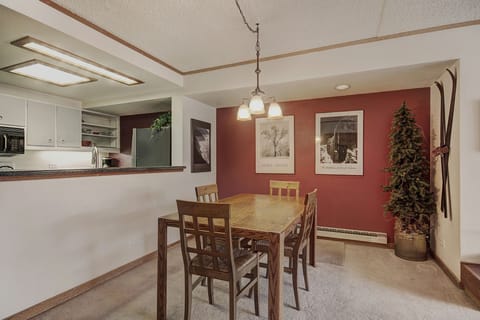 A dining area with a wooden table, chairs, and ceiling light. Red accent wall features three framed pictures. Adjacent kitchen has a pass-through window. Small evergreen tree and decorations in corner.
