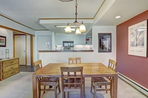 A dining area featuring a wooden table with four chairs. The background shows a kitchen with white cabinets, hanging light fixtures, a framed picture on the right wall, and a wooden dresser on the left.