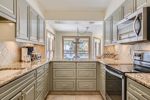 A modern kitchen with olive green cabinets, granite countertops, stainless steel appliances, and a window providing natural light. The room features a U-shaped layout and tiled backsplash.