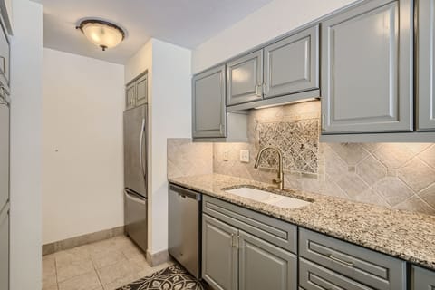A modern kitchen with grey cabinets, granite countertops, stainless steel fridge, under-cabinet lighting, and a tiled backsplash above a white sink with a sleek brass faucet.