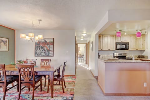 A dining area with a wooden table set for six next to a kitchen. The kitchen has light wood cabinets, granite countertops, and pink pendant lights. There's a hallway leading to other rooms.