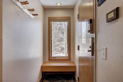 A narrow entryway with a wooden bench under a tall window, a coat rack made from a tree branch on the left wall, and a door on the right side.