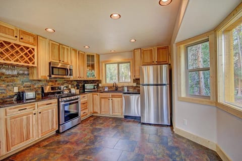A modern kitchen with wooden cabinets, stainless steel appliances, a tiled floor, and large windows overlooking greenery.