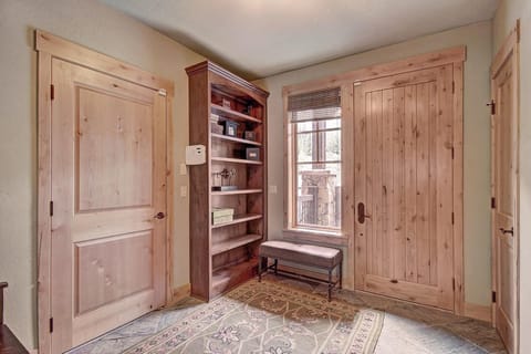 A small room with two wooden doors, a built-in wooden bookcase, a rug on the floor, a window with partial blinds, and a cushioned bench under the window.