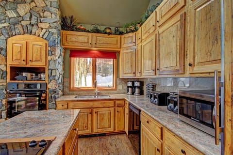 A rustic kitchen with stone and wood features, including wooden cabinets, a granite countertop, various kitchen appliances, and a window with a red valance.