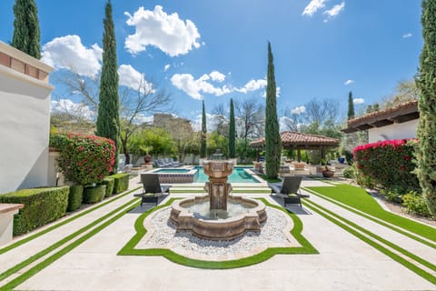 Sunny courtyard view with a central stone fountain, pool, loungers, and shaded outdoor kitchen. Surrounded by tall trees and vibrant landscaping, this space is ideal for relaxing or gathering with your group.