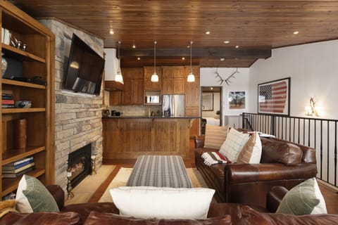 A cozy living room with leather sofas, wooden ceiling, stone fireplace, and a mounted television. The rustic kitchen has wooden cabinets, pendant lights, and a flag wall decor in the background.