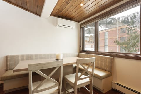 A dining area with a wooden table and two chairs, adjacent to a bench with plaid upholstery, under a wooden ceiling. A large window offers a view of buildings and a snow-covered mountain outside.