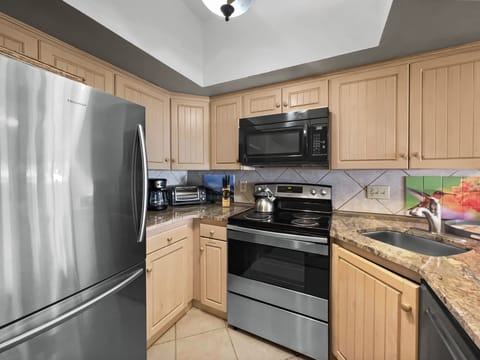 A compact kitchen featuring stainless steel appliances, light wooden cabinets, tiled backsplash and beige countertops, complete with a sink, stovetop, oven, microwave, and small appliances.