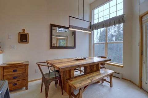 Dining area with a wooden table, benches, and metal chairs under a rectangular light fixture. A large window with a ruffled curtain and a wall mirror reflect natural light into the room.