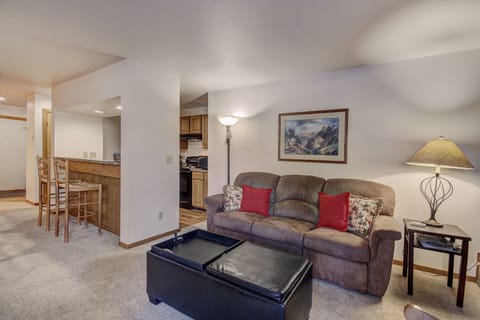 A cozy living room with a brown sofa, red cushions, and a floor lamp. A framed picture hangs above the sofa. A kitchen with wooden cabinets is visible in the background. Two bar stools are at the counter.