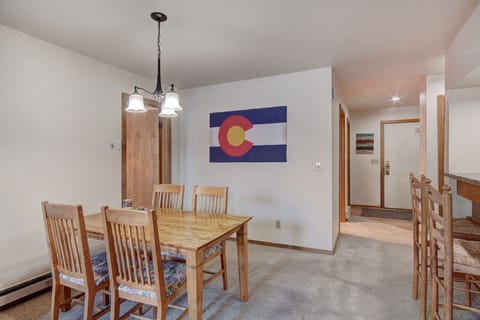 A dining area with a wooden table and chairs, a ceiling light fixture, and a Colorado flag on the wall. There is an open hallway visible in the background.