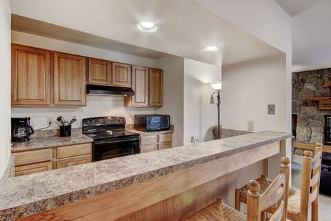 A kitchen with wooden cabinets, a black stove, a microwave, a coffeemaker, and a countertop with two wooden barstools. A stone wall and a fireplace are visible in the background.