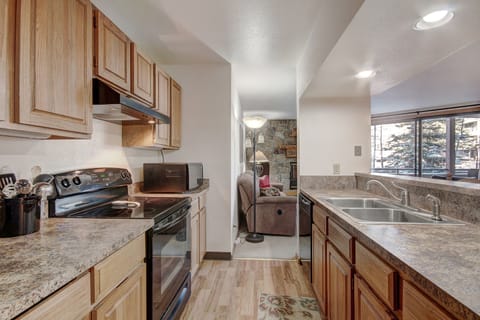 A kitchen with wooden cabinets, a stove, microwave, and sink. An adjacent living area is visible with a person seated on a sofa next to a lamp and window showing a snowy outdoor scene.