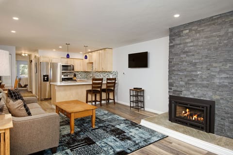 A modern living area featuring a grey sofa, wooden coffee table, and a fireplace with a stone surround. The open kitchen in the background has stainless steel appliances and a breakfast bar with two chairs.