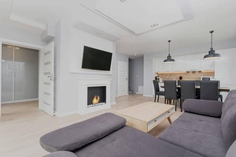 A spacious living room with a cozy sectional sofa, a wooden coffee table, a large wall clock, and abundant natural light coming through the sliding glass doors.