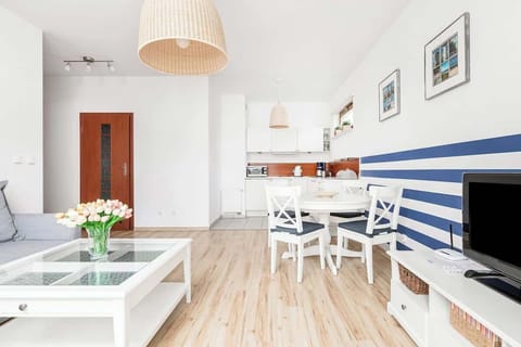 Living room with light-coloured flooring, white furniture and a grey sofa. The dining table is to the right and the open kitchen is visible in the background. The interior is bright and airy, with soft lighting.