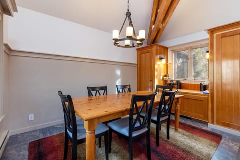 A dining room with a wooden table and six black chairs, a chandelier overhead, and natural light coming through a window. The room has a two-tone wall, exposed wooden beams, and an area rug.