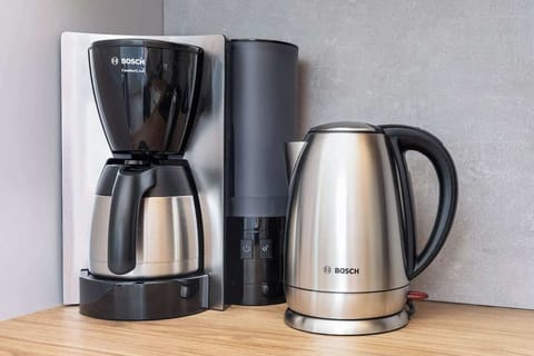 A close-up of a modern coffee station featuring a coffee machine, electric kettle, and cups, placed on a wooden counter for a homey touch.