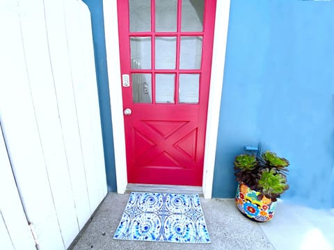 Entrance to Your Home By The Sea has a bold red door, a cute welcome mat, and happy plants that add a cheerful vibe.